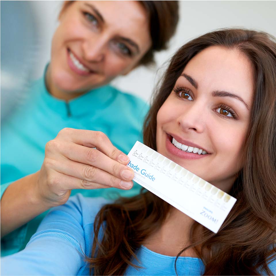 Dental professional holding a shade guide in front of a patient&rsquo;s smile